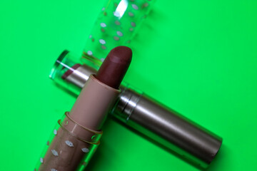 A close-up shows a matte dark red lipstick lying on a bright green background. This contrasting...