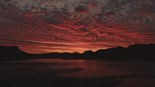 A fiery red and orange sunset over jagged mountain peaks in Iceland. A calm body of water reflects the vibrant sky as the camera slowly pans.