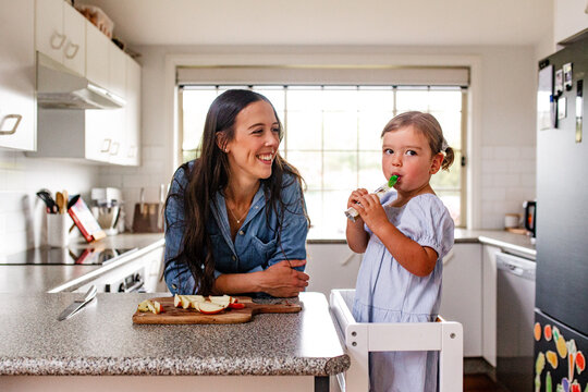 Toddler girl standing on a ladder sipping from food pouch in the kitchen
