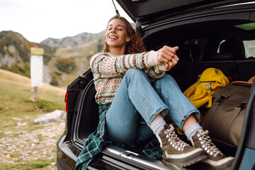 Beautiful traveler with a backpack relaxing in the trunk of a car on a sunny autumn day. Cheerful...