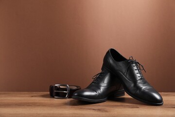 Pair of black leather shoes and belt on wooden surface against brown background, space for text