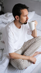 Young brunette man sitting on the edge of a bed, sipping tea while gazing out the window on a...