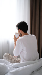 Young brunette man sitting on the edge of a bed, sipping tea while gazing out the window on a peaceful morning