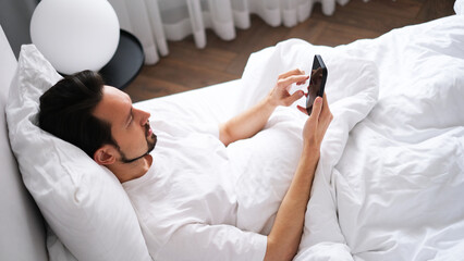 Young brunette man lying in bed looking at his phone early in the morning in a bright cozy bedroom