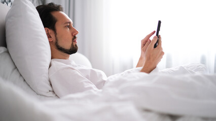 Young brunette man lying in bed looking at his phone early in the morning in a bright cozy bedroom