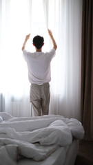 Portrait of a young brunette man opening curtains in a bright cozy bedroom early morning, back view