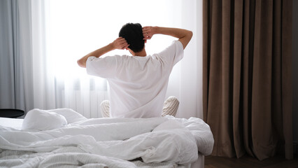 Young brunet man stretching after sleep while sitting on a bed, back view