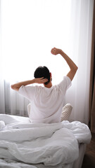 Young brunet man stretching after sleep while sitting on a bed, back view