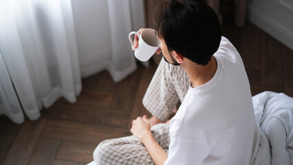 Young brunette man sitting on the edge of a bed, sipping tea while gazing out the window on a peaceful morning