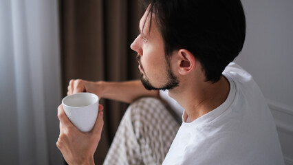 Young brunette man sitting on the edge of a bed, sipping tea while gazing out the window on a peaceful morning