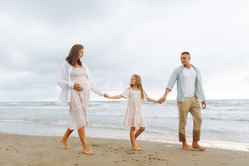 Happy young family walking together on the beach holding hands. Man with his pregnant wife and...