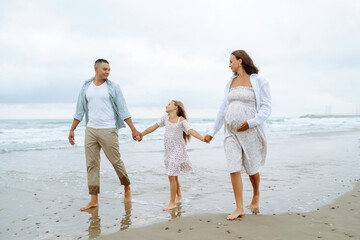 Happy young family walking together on the beach holding hands. Man with his pregnant wife and daughter enjoying time together feeling freedom outdoors. Family, weekend concept.