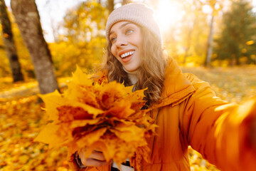 Selfie time! Young woman in bright jacket and bouquet of yellow autumn leaves enjoying sunset in...
