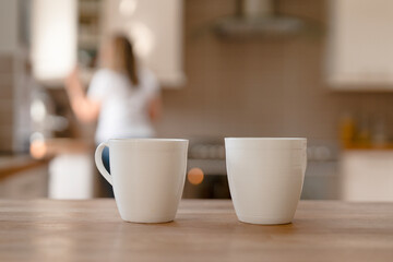 Two white coffee cups on a wooden table with a person preparing food in a modern kitchen during the morning