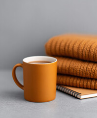 Cozy scene with orange mug filled with warm beverage next to stack of folded sweaters and notebook