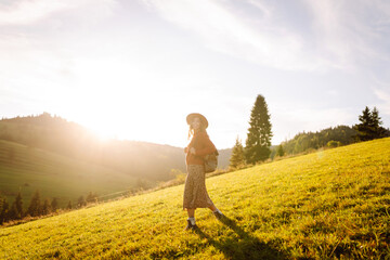 Fototapeta premium Rear view of young female traveler with backpack and hat walking on green hills and enjoying sunset outdoors. Beautiful woman feels freedom against mountain landscape. Hiking and travel concept.