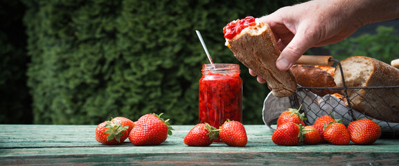 Homemade strawberry jam on a crusty baguette. Breakfast with fresh seasonal fruits outdoors. Horizontal close-up with space for text.