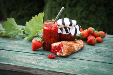 Homemade strawberry jam in a jar with fresh baguette on an old wooden table outdoors. Spread made from fresh seasonal fruit. Close-up.