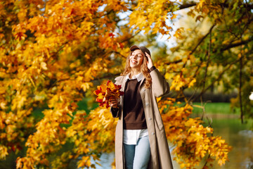 A happy woman in stylish clothes in an autumn park enjoys the sunny weather. Beautiful tourist with...