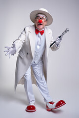 An elderly clown in a white coat, hat and large red patent leather clown boots. Walking stick in...