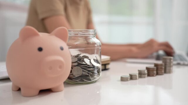 An Asian woman is writing in a notebook and stacking coins and a piggy bank, which symbolizes budgeting, financial planning, saving money and smart money management.