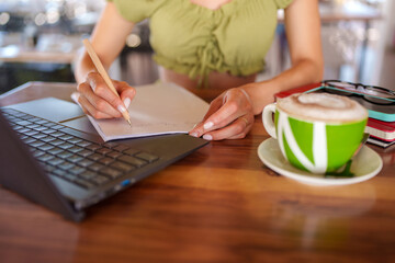 Freelancer writing notes on notepad and working remotely using laptop in a modern bar with cappuccino and smartphone