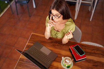 Young woman enjoying a coffee break while working remotely on a laptop, wearing earphones and soaking in the cafe's vibrant atmosphere