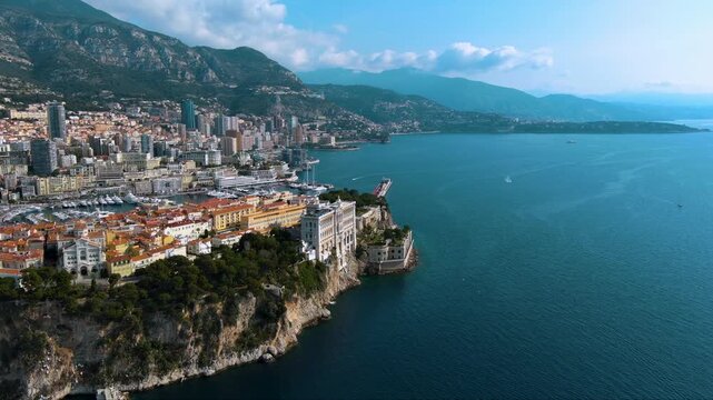 Stunning aerial flight revealing the Oceanographic Museum on the Rock of Monaco, Port Hercules, and the Monte Carlo cityscape against the Mediterranean Sea and mountains.
