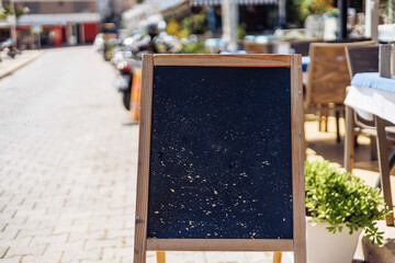 A wooden chalkboard sign stands on a sunny street. The background features outdoor seating and a bustling atmosphere. The scene is vibrant and inviting.