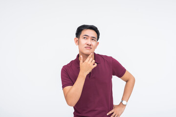 A pensive young Asian man with one hand on his chin and the other on his waist, looking up in deep...