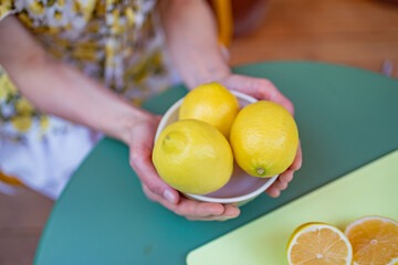 A woman is holding a white bowl filled with fresh, vibrant lemons on a green table, with halved lemons visible in the background, emphasizing a healthy and fresh lifestyle
