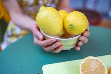 A woman holds a bowl of fresh, vibrant lemons on a green table, with a sliced lemon visible on a yellow cutting board, emphasizing freshness and culinary preparation