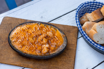 Sizzling shrimp dish served in a black skillet on a wooden board. Accompanied by a basket of fresh bread rolls. Ideal for food lovers and culinary enthusiasts.