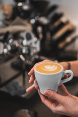 Barista holding a white cup of coffee with heart-shaped latte art in a cozy café setting, with a blurred coffee machine in the background