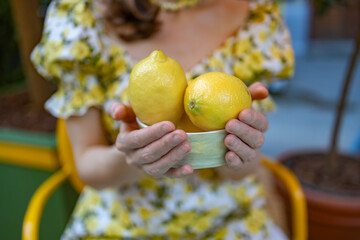 A woman in a floral dress holds a white bowl filled with fresh, vibrant lemons, surrounded by a cozy outdoor atmosphere with potted plants and a wooden table