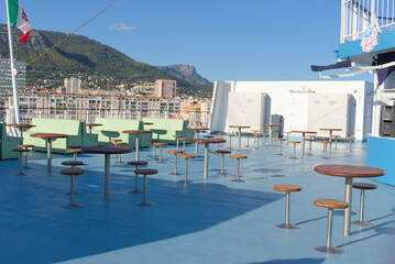 Fototapeta premium An outdoor deck of a ship features numerous high-top tables and stools, green seating, and an Italian flag waving, with a coastal town and mountains in the background under a clear sky.