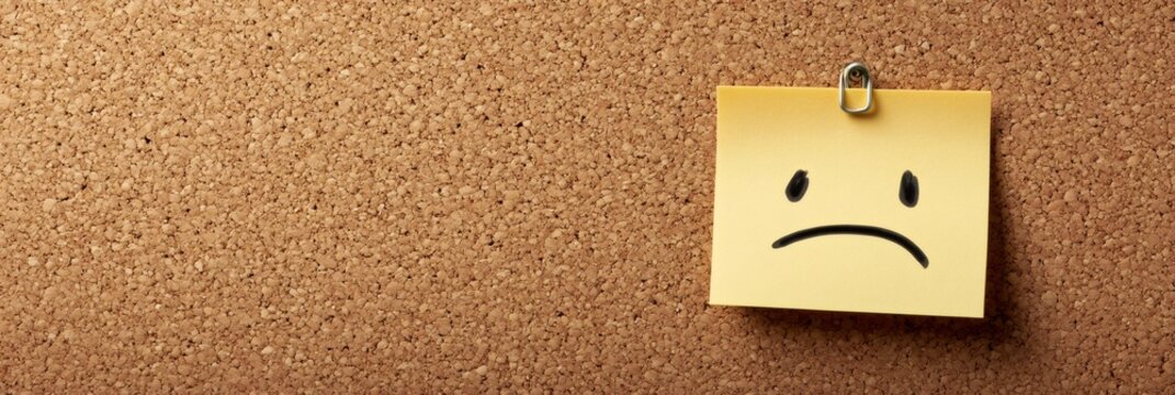 Yellow sticky note with sad face hanging on a cork board representing concepts such as stress, stress relief, anger management, deadline, time management, and therapy