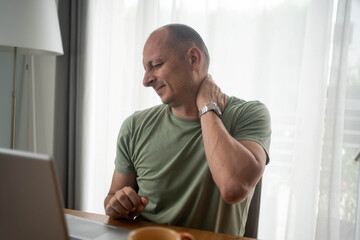 Mature man feeling neck pain while working from home with laptop, suffering from tension,...