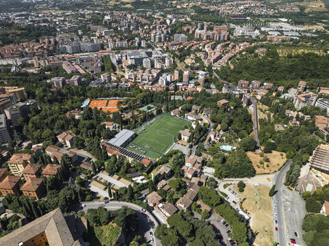 Aerial view of terracotta rooftops cascade down gentle slopes, punctuated by lush green trees and the bright rectangle of a football field, Perugia, Umbria, Italy.