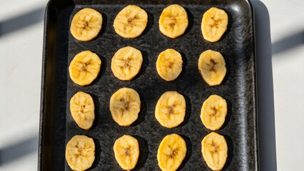 Dehydrated banana chips slices arranged neatly on a dark rectangular baking sheet