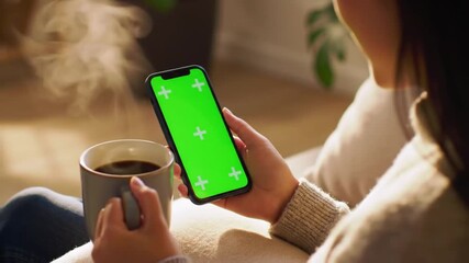 An individual with a smartphone that has a green mock-up screen in upright mode, drinking coffee. The man is navigating the Internet and consuming content, including videos - Powered by Adobe