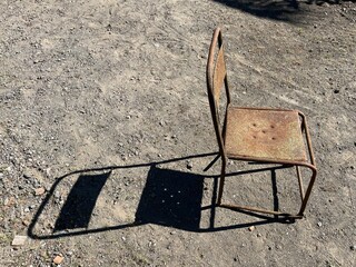 Old rusty metal chair standing alone on dry gravel ground in sunlight, showing weathered texture and aged condition.