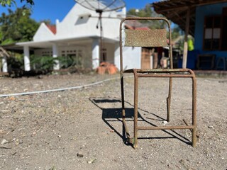Rusty metal chair with missing seat placed on dry ground in front of old houses, showing signs of decay and abandonment.