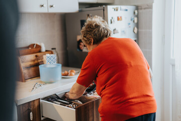 An elderly woman in a bright orange shirt organizes kitchen utensils in a drawer, demonstrating...