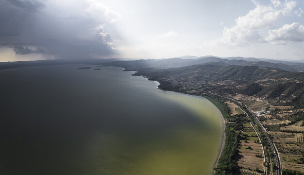 Aerial view of the vast, calm lake mirroring the sky's drama, where the shoreline meets the rolling hills dotted with buildings, Trasimeno Lake, Torricella, Umbria, Italy.