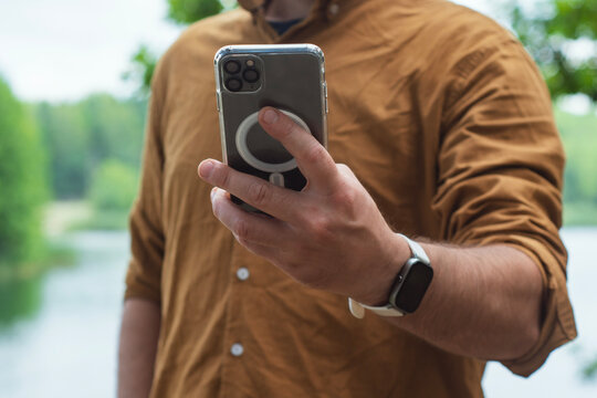 Close-up of a man using a smartphone with a transparent case, standing outdoors near a lake. Concept modern digital lifestyle, mobile connectivity on the go in a natural environment.