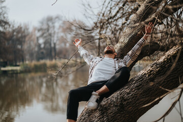A man enjoys a relaxed moment draped over a tree branch near a tranquil river, surrounded by a...