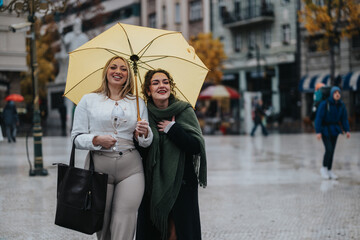 Smiling friends standing closely under a yellow umbrella in an urban setting on a rainy day, expressing togetherness and joy despite overcast skies and light rain. The background features a city
