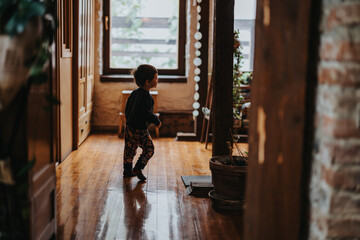 A young child walking inside a cozy house with rustic wooden interiors and large windows, creating...
