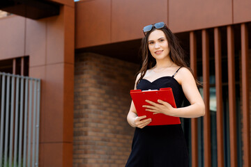 Young woman in black dress holding red tablet, smiling and looking at camera. Brick wall and metal...